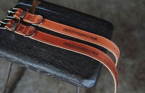 Zwei braune Lederhalsbänder mit Metallschnallen, auf denen „HERZENSBRECHER“ eingraviert ist, auf einer dunklen strukturierten Oberfläche.