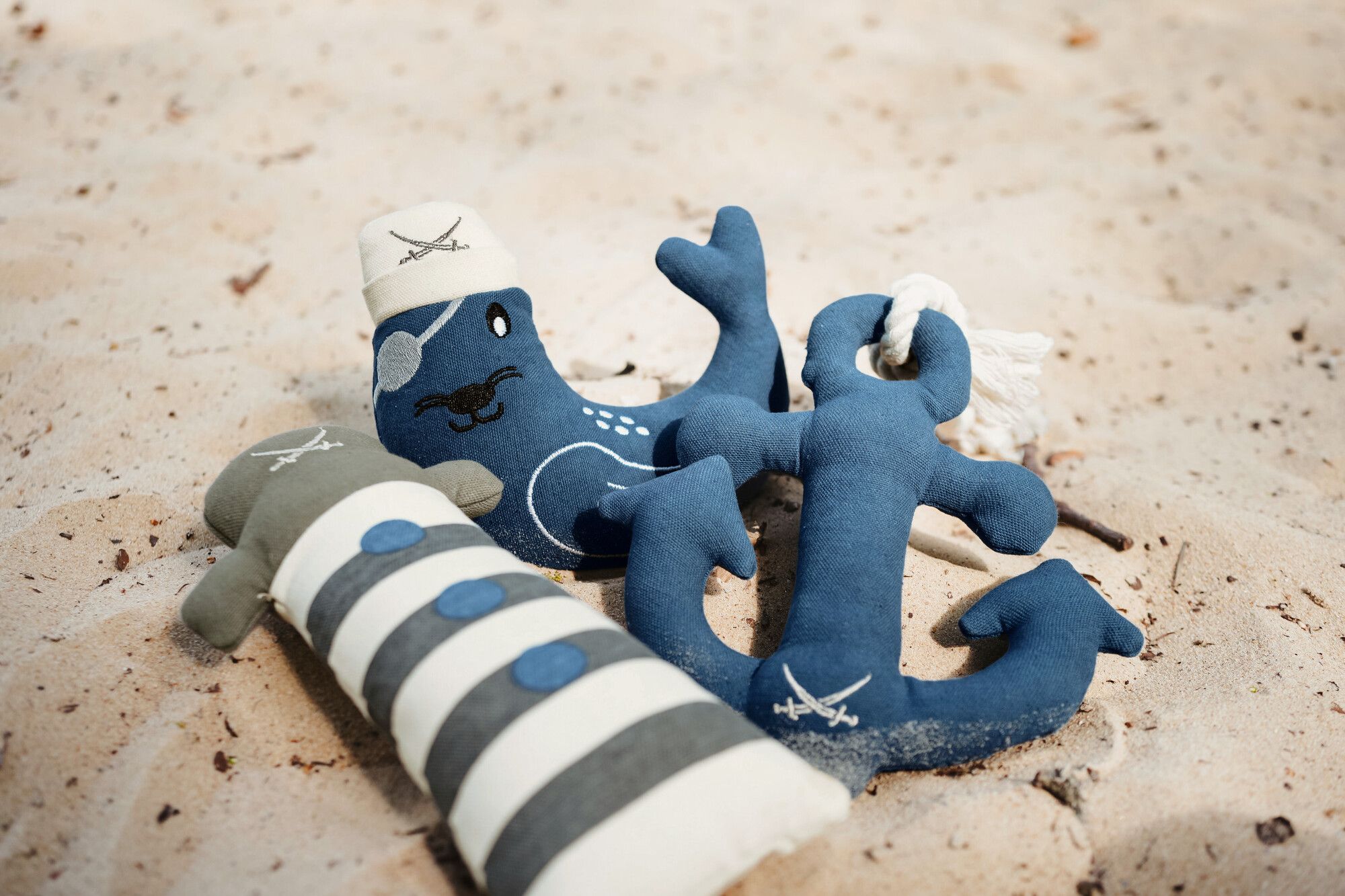 Plüschtiere im maritimen Stil auf dem Sand: ein blauer Anker, ein gestreifter Leuchtturm und eine blaue Robbe mit einer Matrosenmütze.