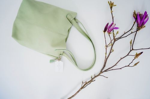 Eine hellgrüne Tragetasche mit einem weißen Etikett, platziert neben einem Zweig mit violetten Blüten vor einem weißen Hintergrund.