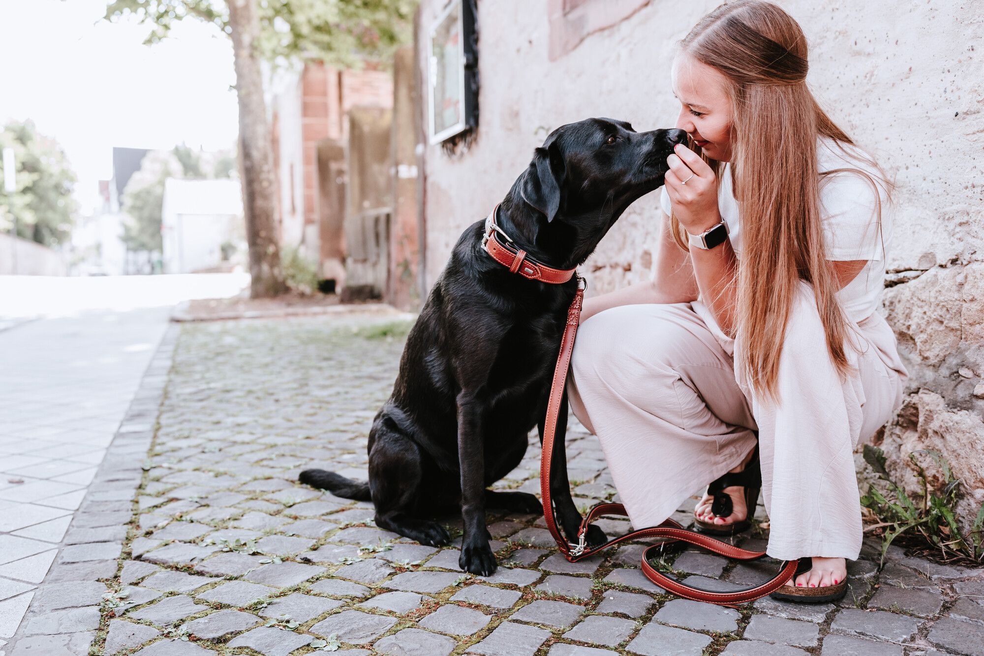 Eine Frau hockt auf einem Kopfsteinpflasterweg und lächelt einen schwarzen Hund an. Der Hund leckt ihr spielerisch das Gesicht.