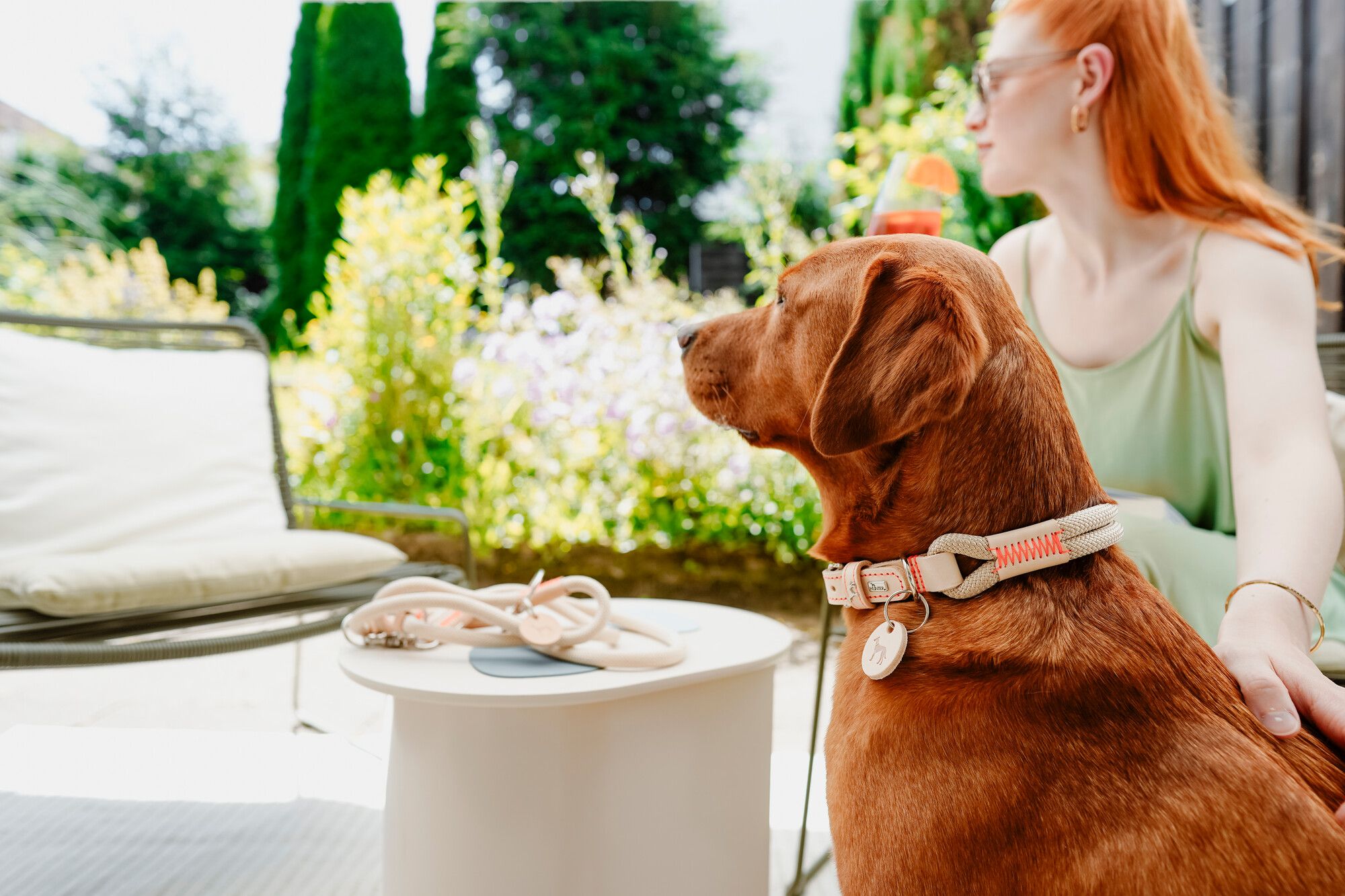 Eine Frau mit roten Haaren sitzt in einem Garten und hält sanft einen braunen Hund neben sich. Vor ihnen auf dem Tisch liegt eine Leine.