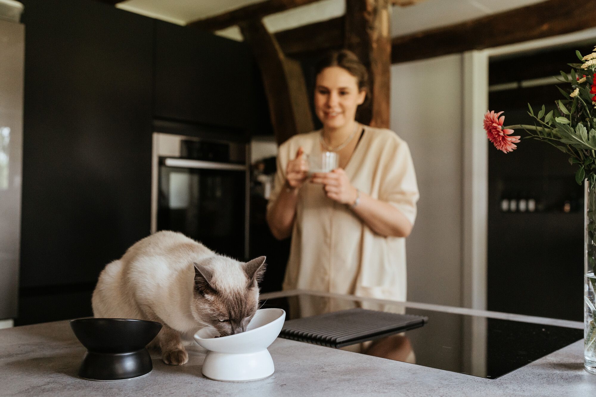 Eine Katze trinkt aus einem weißen Napf auf einer Küchenarbeitsplatte, während im Hintergrund eine Frau mit einer Tasse in der Hand neben einem schwarzen Schrank und Blumen steht.