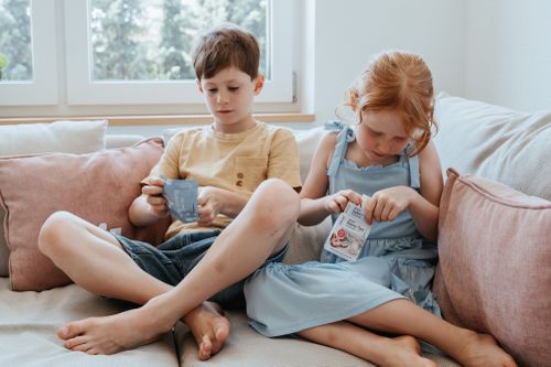 Zwei Kinder sitzen auf einem Sofa und sind ganz damit beschäftigt, Mysteryboxen zu öffnen; um sie herum liegen weiche Kissen, im Hintergrund ist ein Fenster zu sehen.