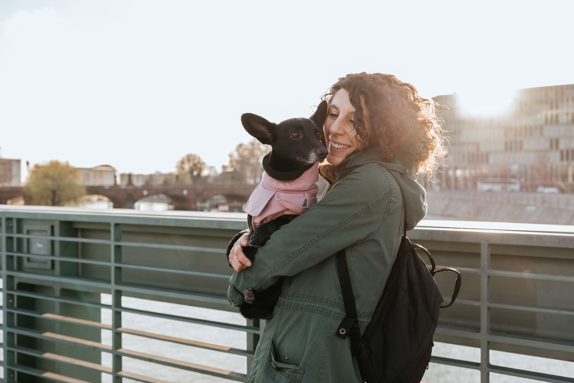 A person with curly hair smiles while holding a small black dog in a pink jacket on a bridge, with a sunny cityscape in the background.