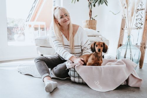 Eine Frau sitzt drinnen mit einem kleinen Hund neben ihr in einem Korb auf einer Decke. Umgeben von einer gemütlichen Einrichtung. Helles, natürliches Licht, das durch ein Fenster fällt, unterstreicht die friedliche Atmosphäre.