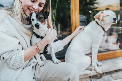 Eine Frau sitzt im Freien und lächelt, während sie einen kleinen schwarz-weißen Hund im Arm hält; ein weiterer weißer Hund sitzt neben ihr.
