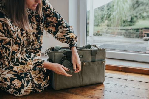 Eine Person in einem geblümten Kleid sitzt am Fenster und befestigt eine kleine Tasche an einer grünen Stofftasche auf einem Holzboden.