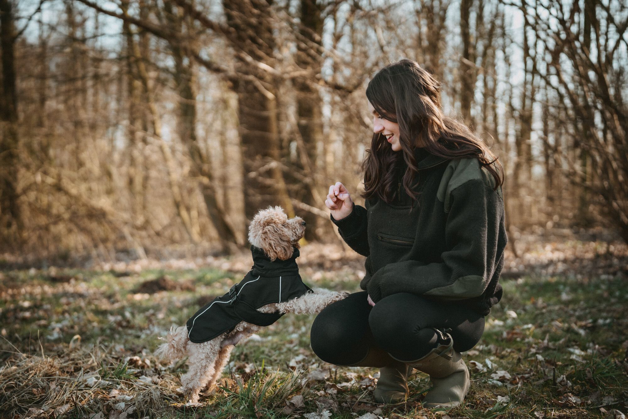 A woman kneels on grass, smiling and holding a treat for a small dog in a jacket, with trees in the background.