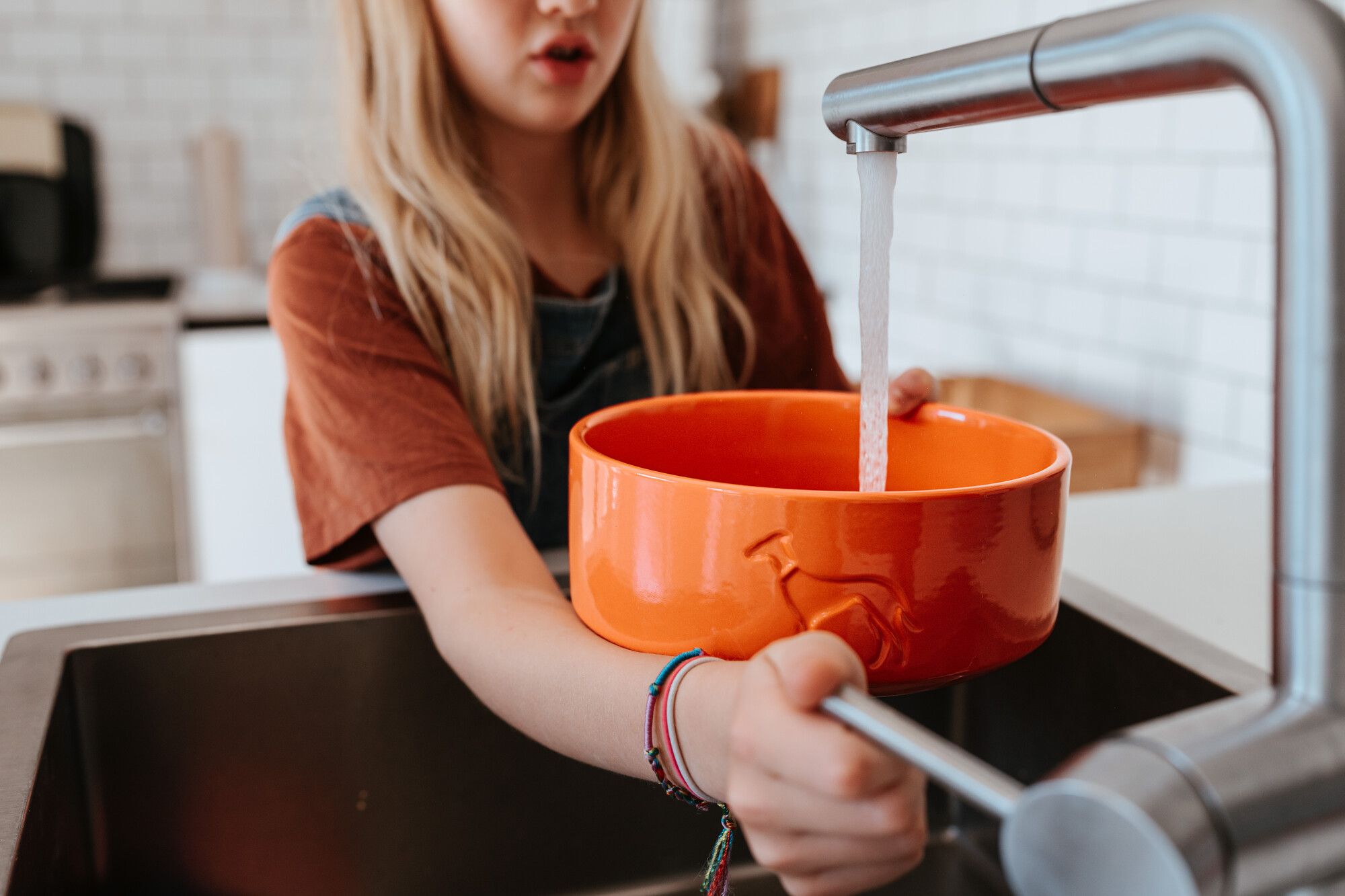 Eine Person füllt eine orangefarbene Schüssel mit Wasser aus einem Küchenwasserhahn. Die Schüssel ist mit einem Hundesilhouette-Design verziert.