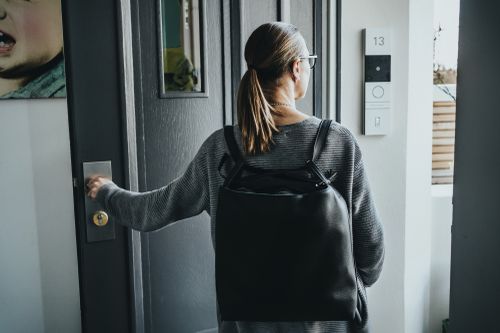 Eine Frau mit Pferdeschwanz und Brille, die einen grauen Pullover und einen schwarzen Rucksack trägt, öffnet eine Tür in einem modernen Flur.