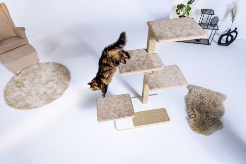 Langhaarige Katze springt dynamisch von einer der gepolsterten Plattformen eines modernen Kratzbaums; helle, minimalistische Wohnraumszene mit Teppich, Sessel und Dekoration im Hintergrund.