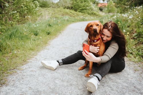 Frau sitzt auf einem Weg, umarmt einen braunen Hund und macht dabei ein Selfie mit einem roten Telefon, umgeben von viel Grün.