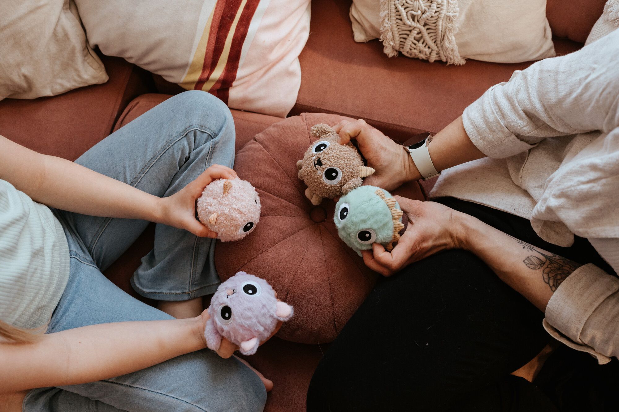 Zwei Menschen sitzen auf einem Sofa, halten kleine, bunte Plüschtiere mit großen Augen in den Händen und sind von Kissen umgeben.