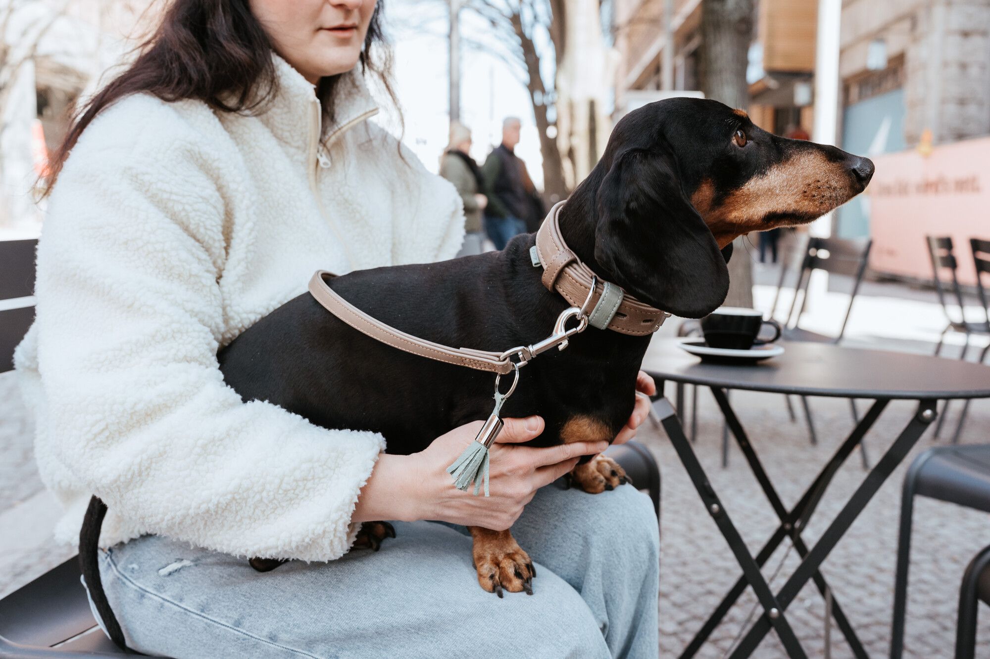 Eine Person in einem weißen Pullover sitzt in einem Straßencafé, auf ihrem Schoß ein schwarz-brauner Dackel.