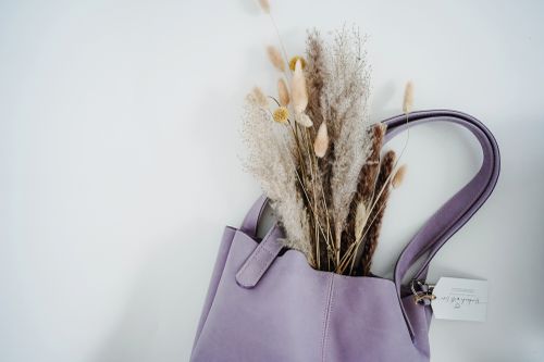 Eine lavendelfarbene Handtasche mit getrockneten Blumen, darunter Pampasgras und gelbe Blüten, die vor einem weißen Hintergrund hervorblitzen.