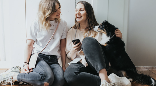 Zwei Frauen sitzen lächelnd auf dem Boden, eine von ihnen hält ein Telefon in der Hand. Ein schwarz-weißer Hund sitzt neben ihnen und schaut sie liebevoll an.
