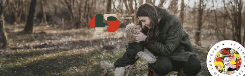 Eine Frau kniet in einem Wald nieder, um einen kleinen Hund zu umarmen. Die Szene wirkt ruhig, im Hintergrund sind kahle Bäume zu sehen. Das Bild ist mit Logos überlagert.