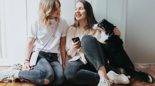Zwei Frauen sitzen auf dem Boden, lächeln und unterhalten sich. Eine hält ein Telefon in der Hand, während ein schwarz-weißer Hund neben ihnen sitzt.