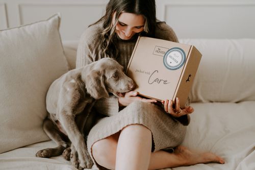 Eine Frau sitzt auf einem Sofa, lächelt einen Welpen an und hält dabei ein Kartonpaket mit der Aufschrift „packed with Care“ in der Hand.
