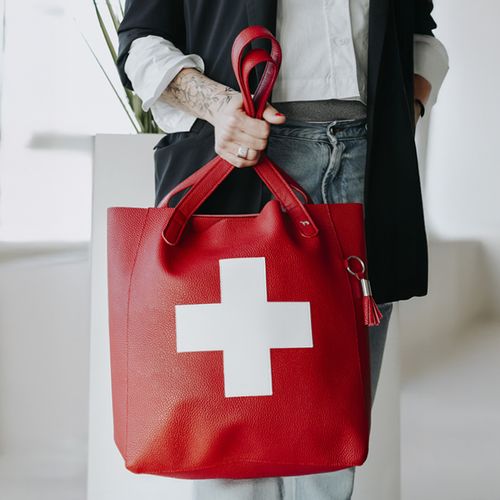 Person mit einer roten Tragetasche mit weißem Kreuzmuster, bekleidet mit schwarzer Jacke und Jeans, stehend in einem Innenraum.