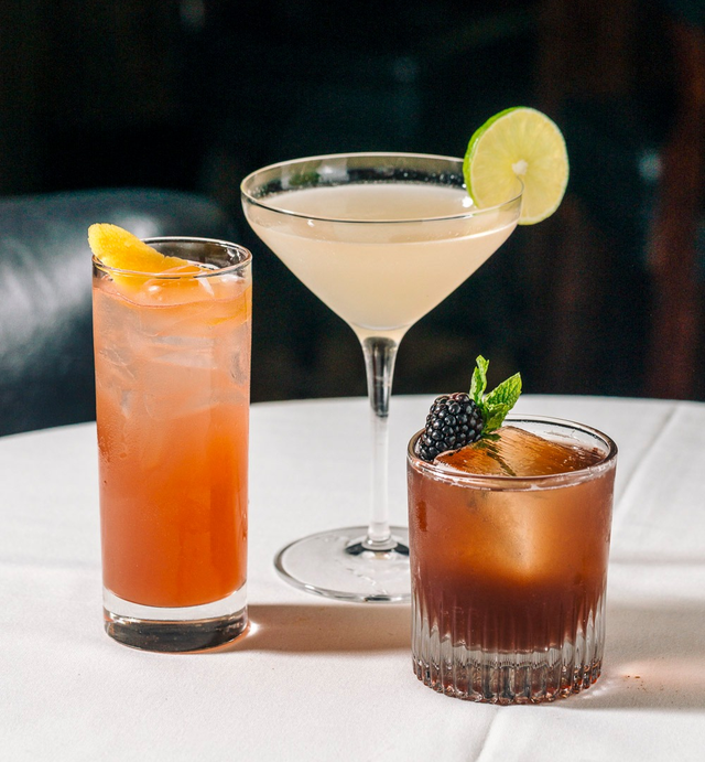 Three cocktails on a table: a tall iced drink with an orange slice, a martini with a lime wedge, and a whiskey glass with a mint sprig and blackberry.