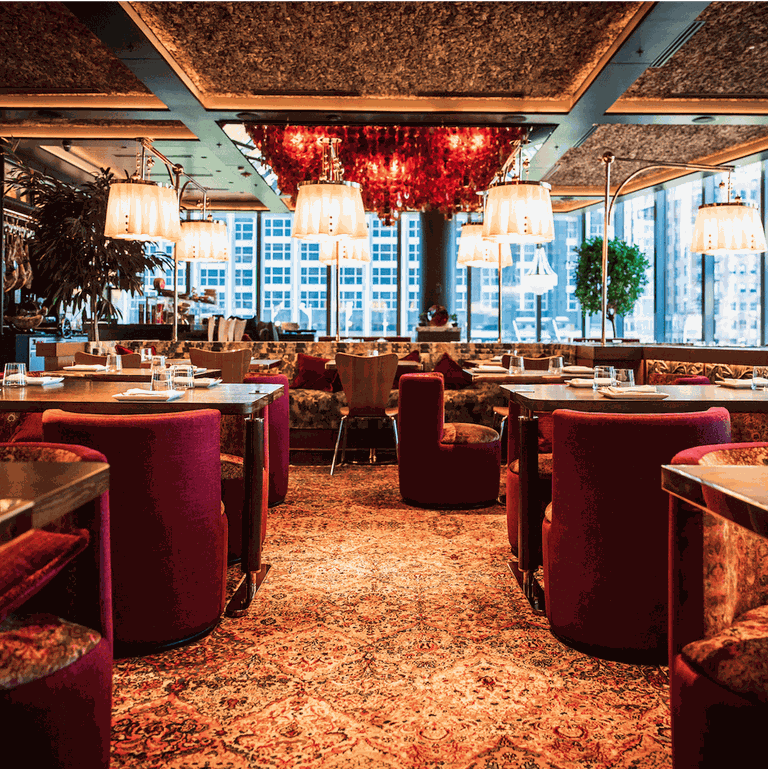 Elegant restaurant interior with red chairs, patterned carpet, and hanging lamps. Large windows reveal a Chicago cityscape in the background.