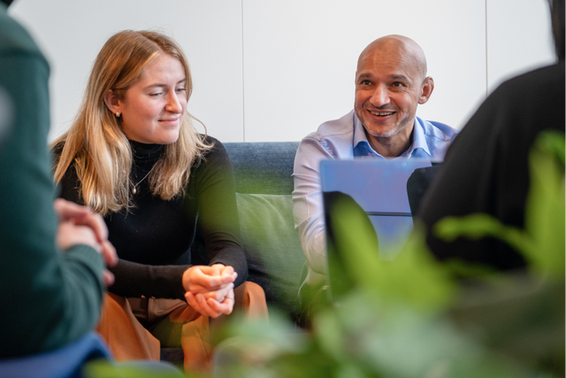 Two people sitting on a couch in an office, one looking at a computer and the other engaging in conversation with someone else outside of the shot