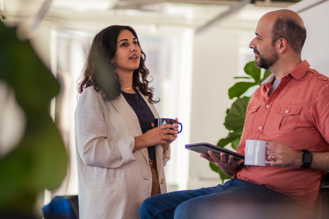 A photo of two people in an office talking casually over cups of coffee