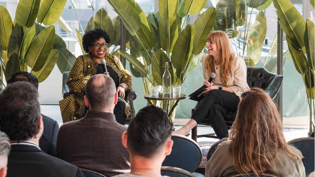 Two women sit and converse on stage with microphones, surrounded by large green plants, while an audience listens attentively in the foreground.