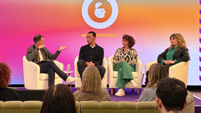 Four people sit on stage in a panel discussion with a colorful backdrop, engaging with a seated audience.