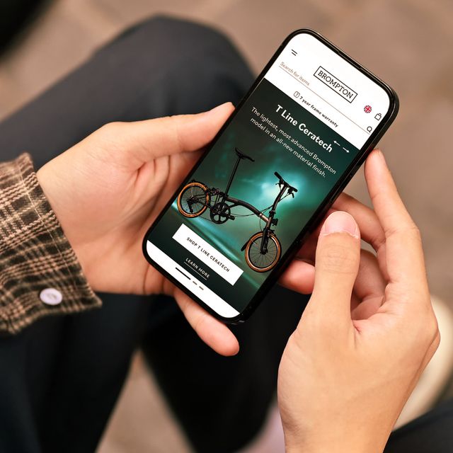 Person holding a smartphone displaying the Brompton website, featuring a black folding bike with orange wheels on the screen.