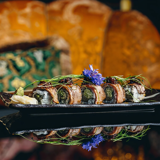 Sushi rolls topped with grilled fish and garnished with edible flowers and greens on a black plate, set against a colorful, ornate background.