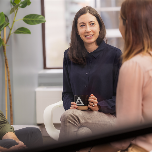 A woman wears a blue button up shirt and plaid pants, she holds an Apply Digital mug. She talks to another woman wearing a pink blouse who appears blurred in the foreground.