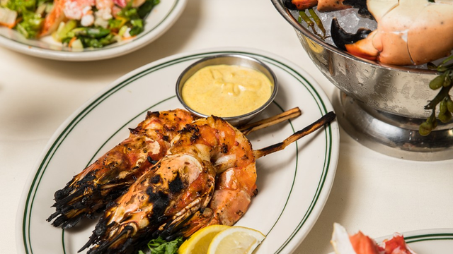Grilled shrimp skewers on a plate with lemon wedges and sauce, alongside a seafood salad and crab dish in a metal bowl.