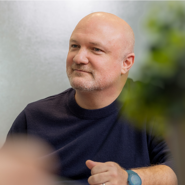 Gavin wears a dark blue sweater. He looks off into the distance. In the foreground are some blurred plants.