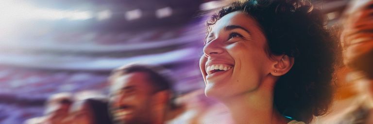 A sports fan with curly hair smiles joyfully in a blurred, colorful stadium setting, capturing a moment of excitement.