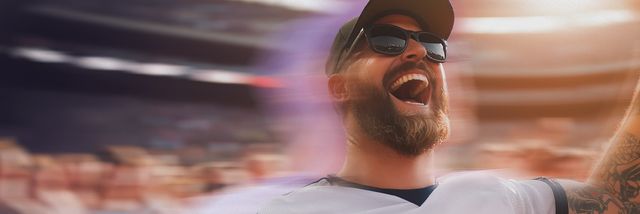 A bearded man wearing sunglasses and a cap cheers energetically at a sports event, with a blurred crowd and stadium in the background.