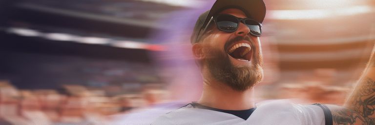 A bearded man wearing sunglasses and a cap cheers energetically at a sports event, with a blurred crowd and stadium in the background.