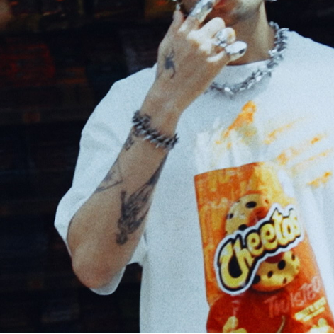 Person holding a bag of Cheetos, wearing a chunky chain necklace, holding fingers near mouth, with tattoos visible on arms.