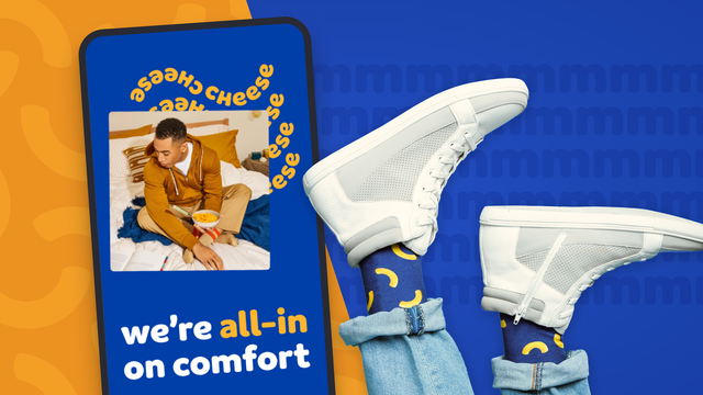 A pair of legs are in the air, wearing white shoes and blue socks with yellow macaronis on them. A mobile screen shows the Kraft Mac & Cheese site.