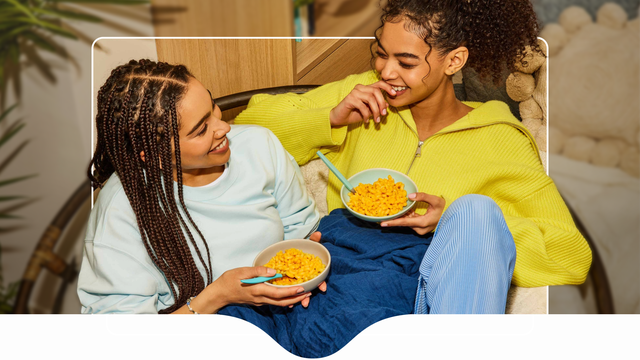 Two women eat Kraft Mac'n'Cheese while sitting in the same chair.