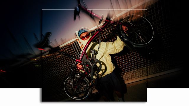 Person in a yellow jacket and helmet holding a bicycle upright at night, with a blurred background and a metal fence behind.