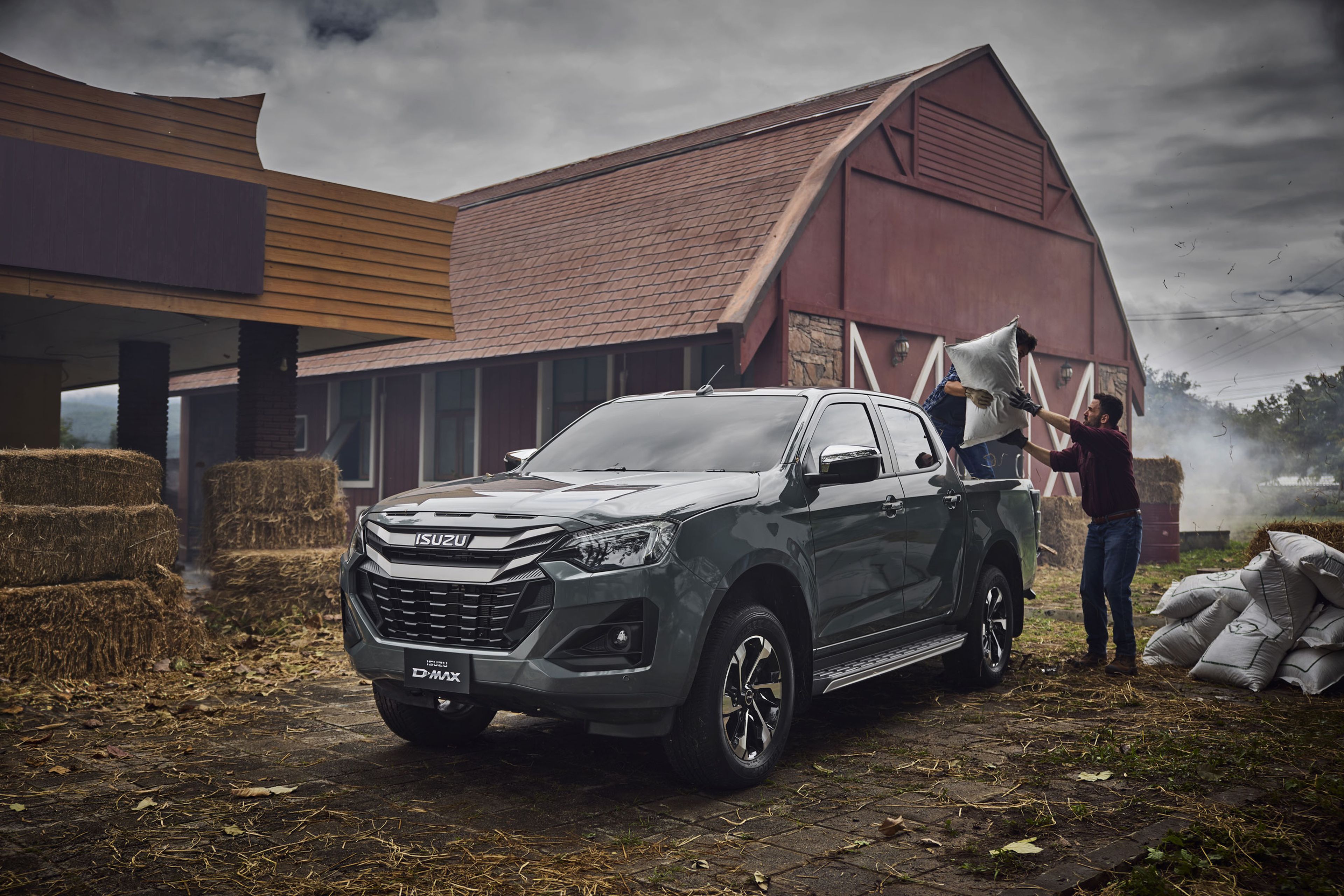 A green ISUZU D-MAX parked near a barn, with two people loading large bags into the truck. Hay bales are stacked nearby.