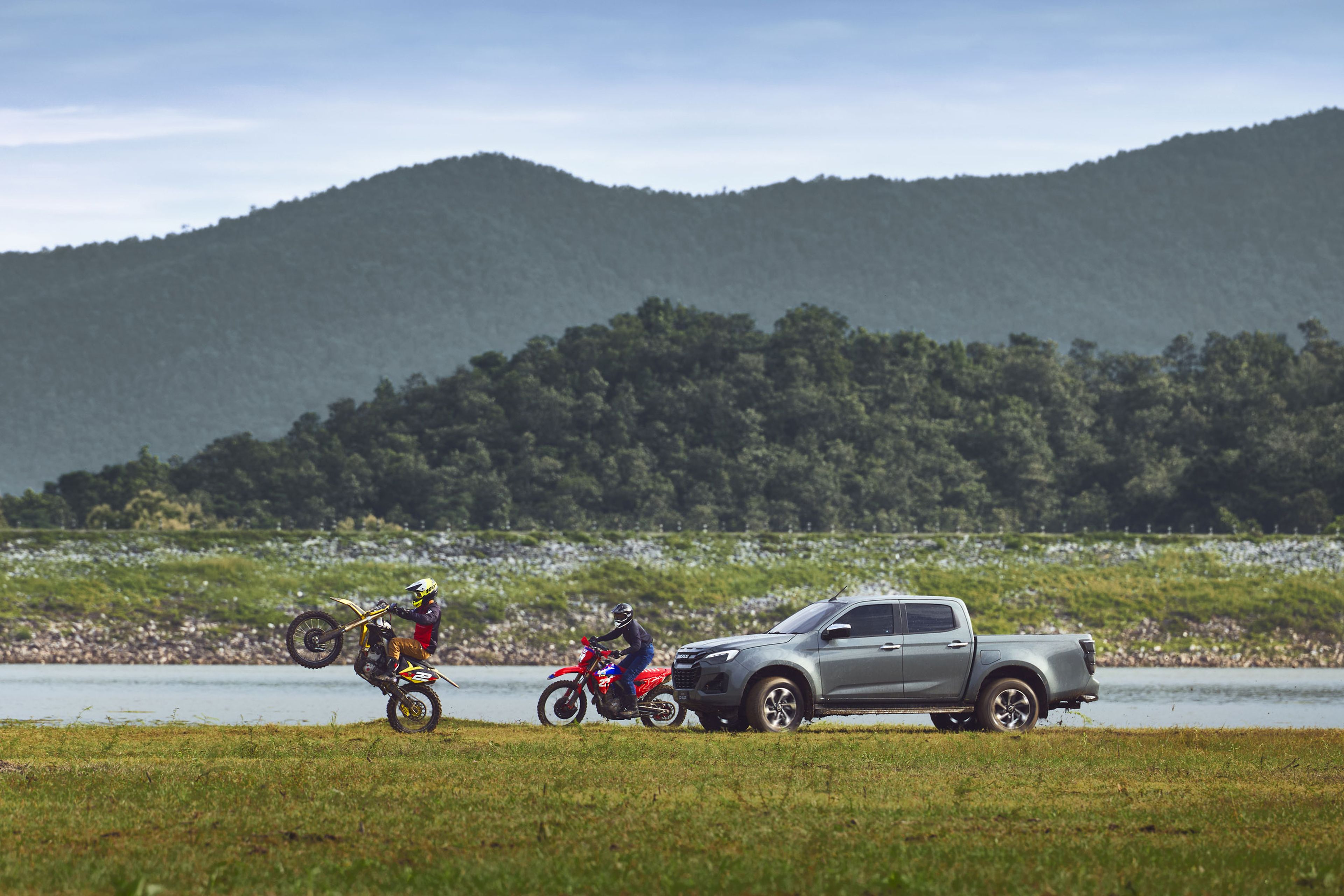 Two motorbikes and an ISUZU D-MAX near a lake, with a rider performing a wheelie. Hills and a cloudy sky form the backdrop.