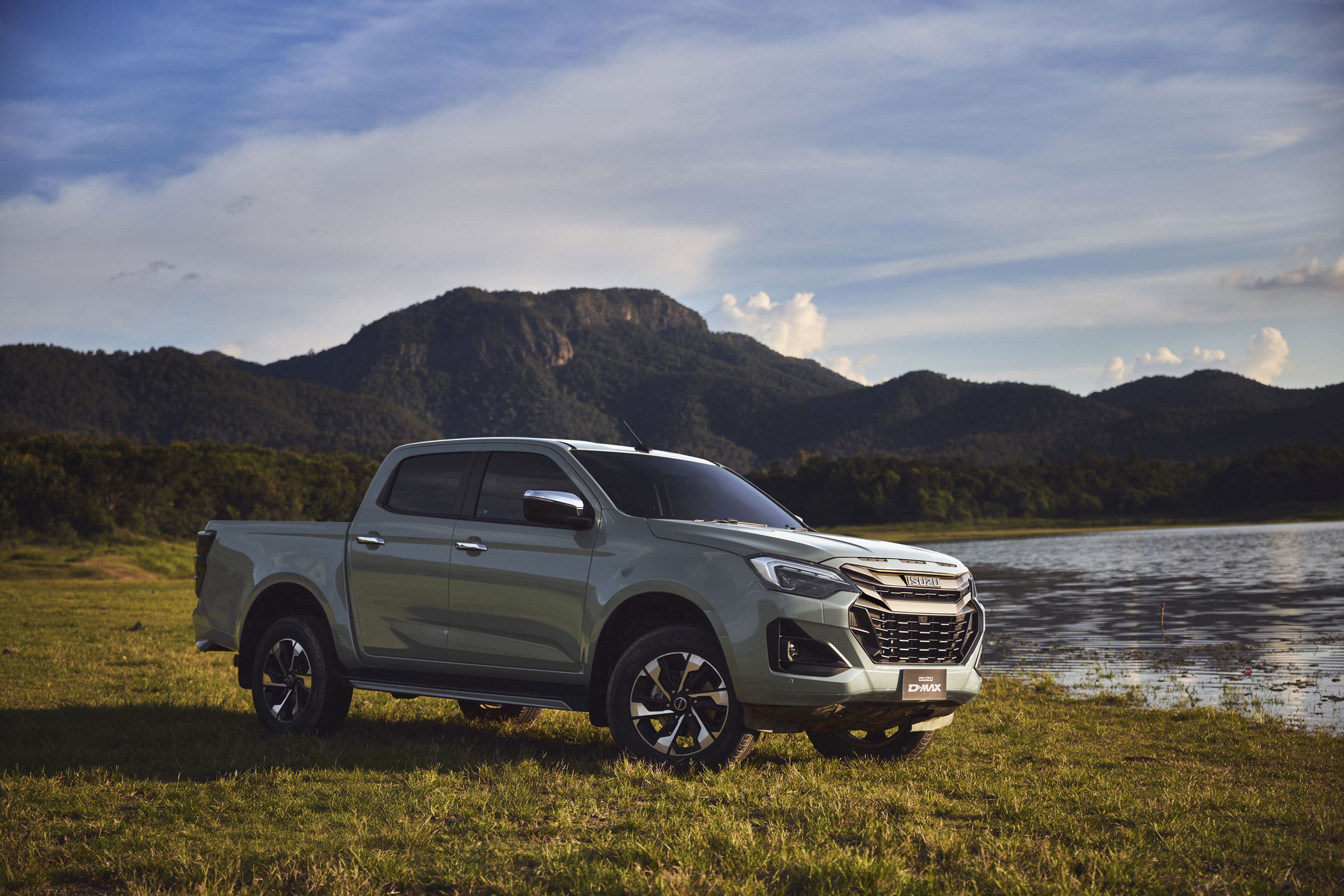 An ISUZU D-MAX parked on a grassy field by a lake, with mountains and a blue sky in the background.