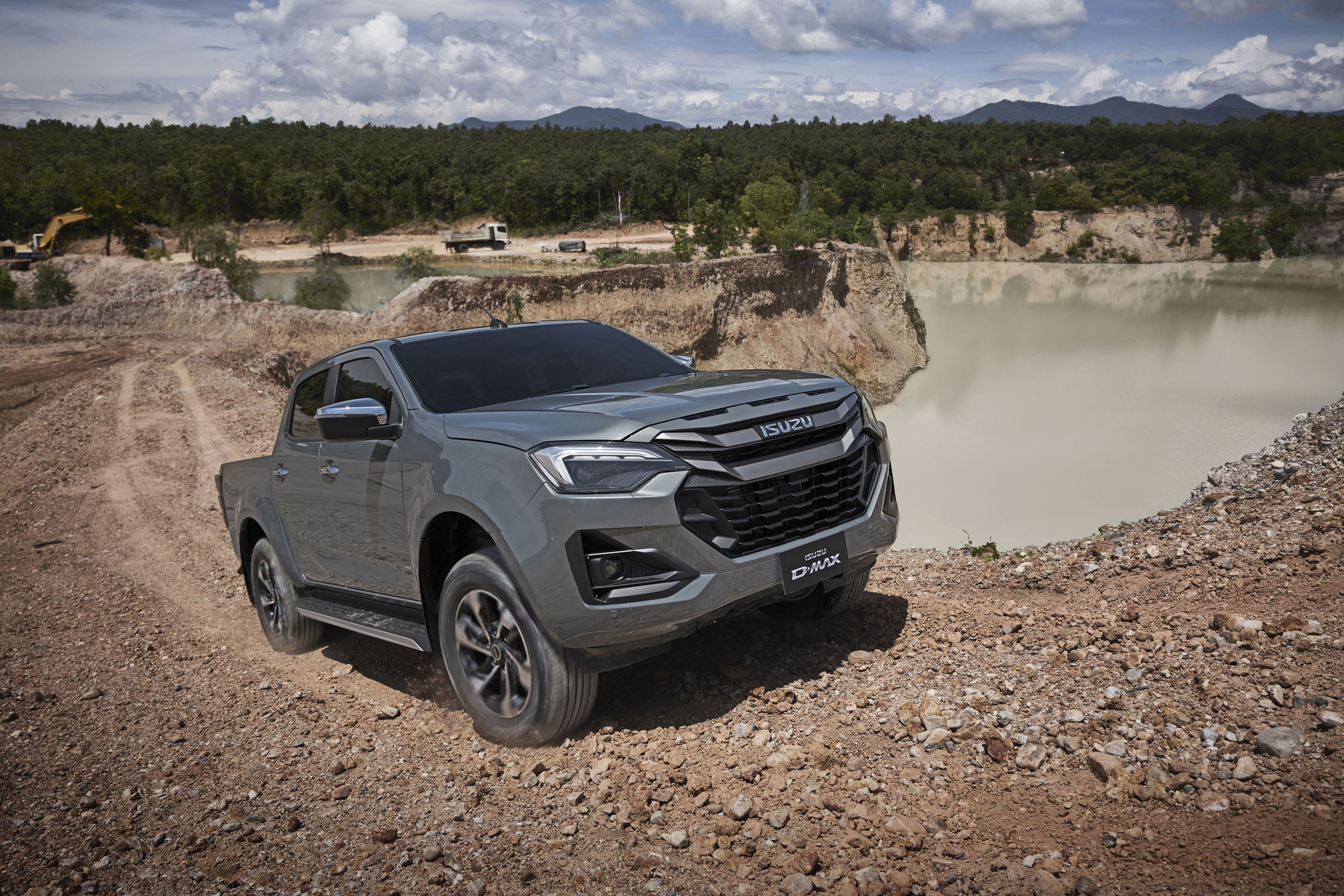 Gray ISUZU D-MAX navigating rough terrain near a water body, with a forest and cloudy sky in the background.