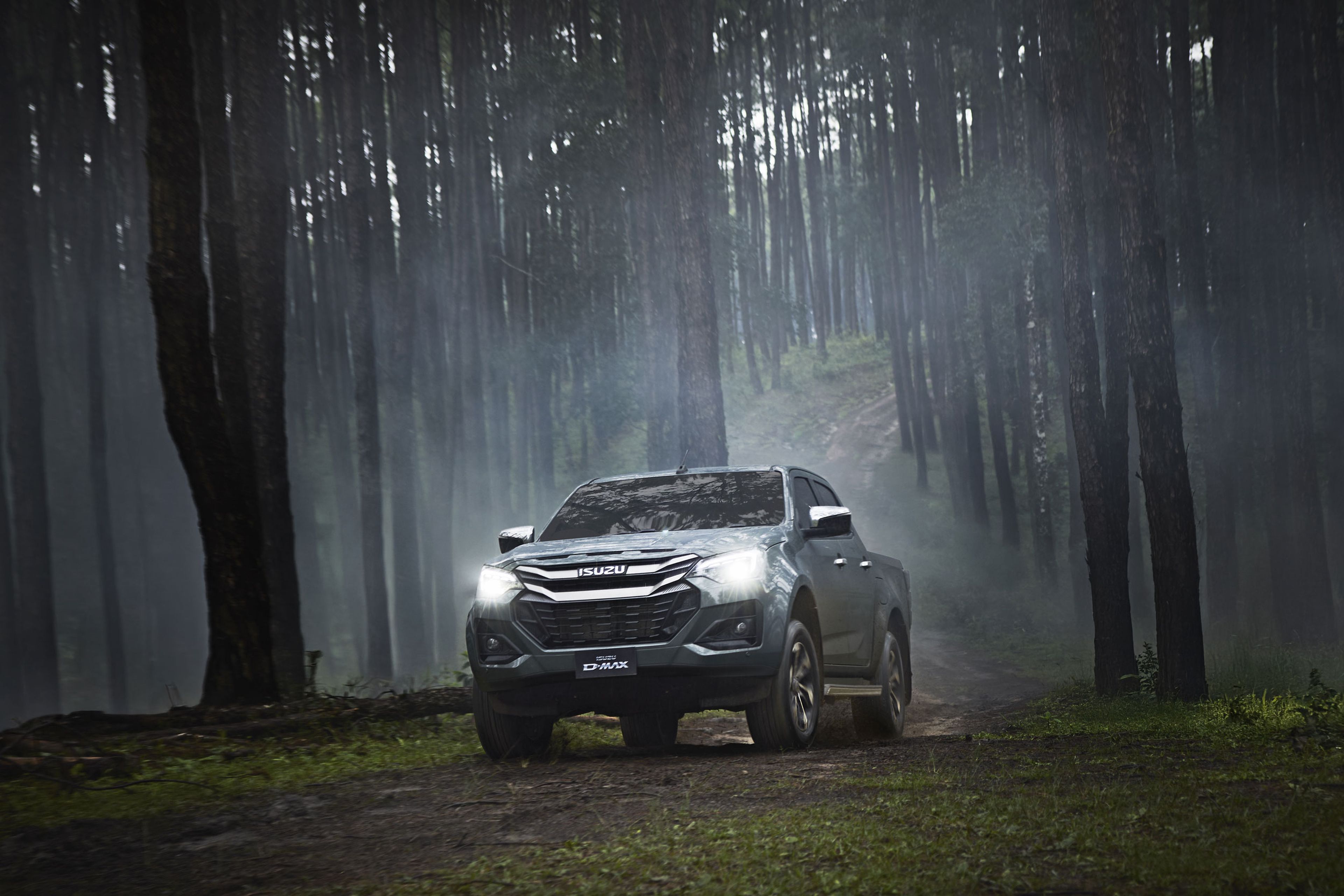 An ISUZU D-MAX with headlights on navigates a foggy, dense forest path lined with tall trees.