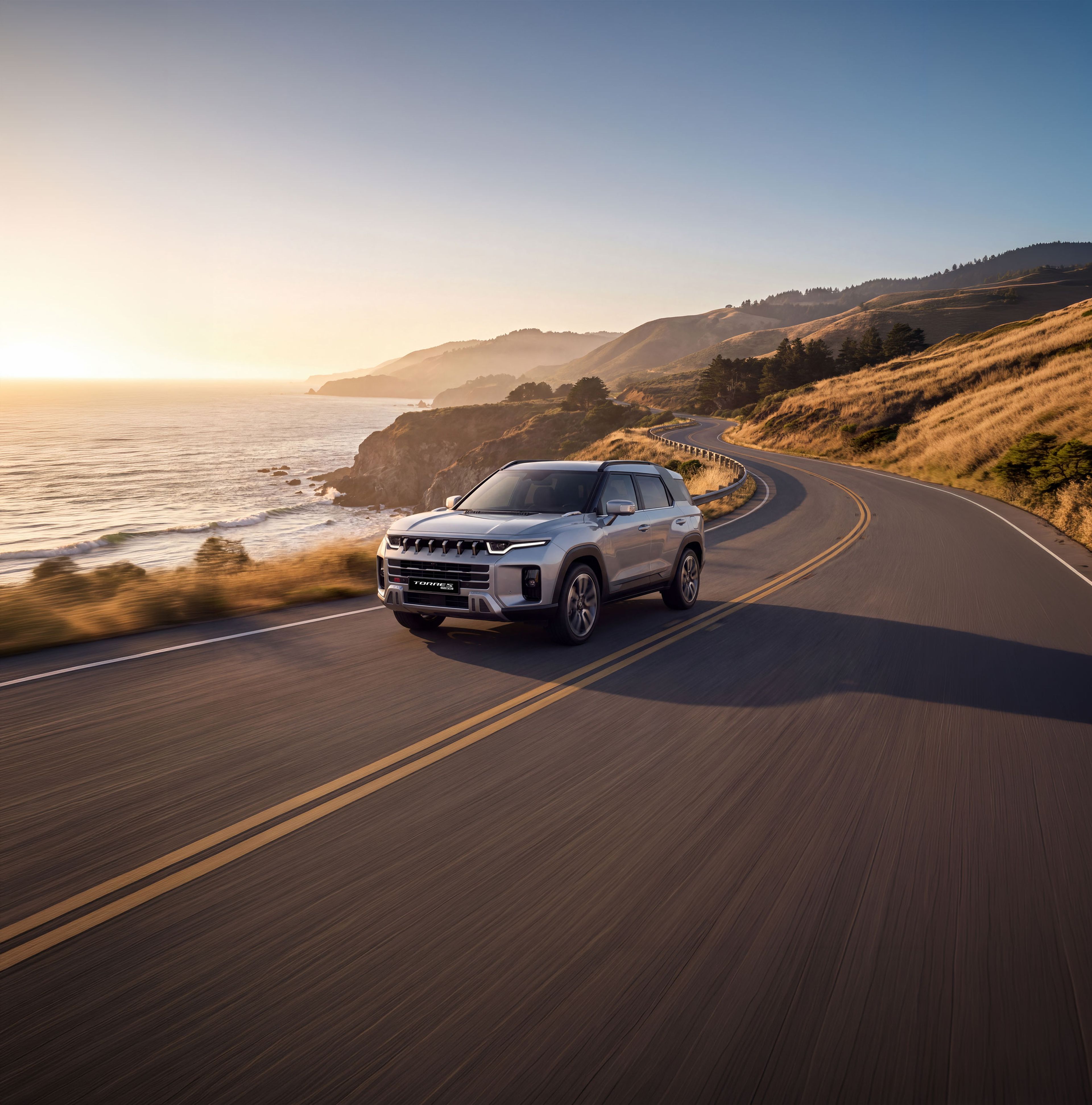 A silver KGM Torres Hybrid driving on a scenic coastal road at sunset, with cliffs and the ocean in the background.
