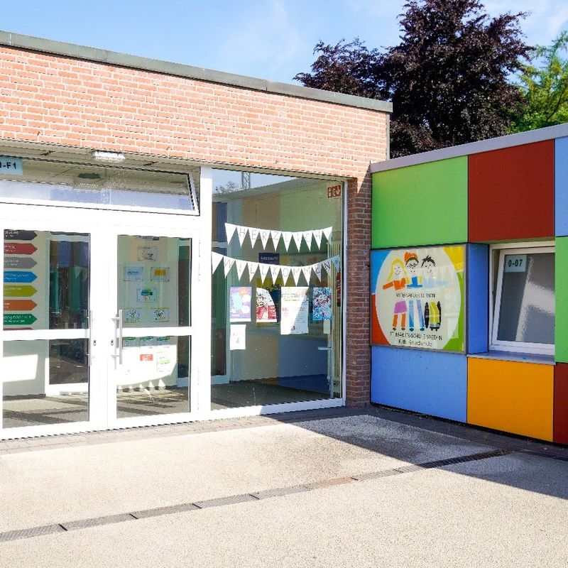 Bild des Eingangs der St. Marien-Schule. Links ist die Glasfront mitsamt der Eingangstür zu sehen. Rechts befindet sich eine farbige Seitenwand und das Logo der Schule