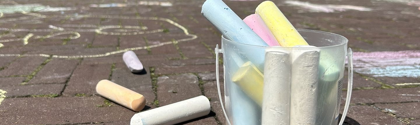 Becher voller bunter Kreide auf bemalten Marktplatz. Im Hintergrund malen Kinder.